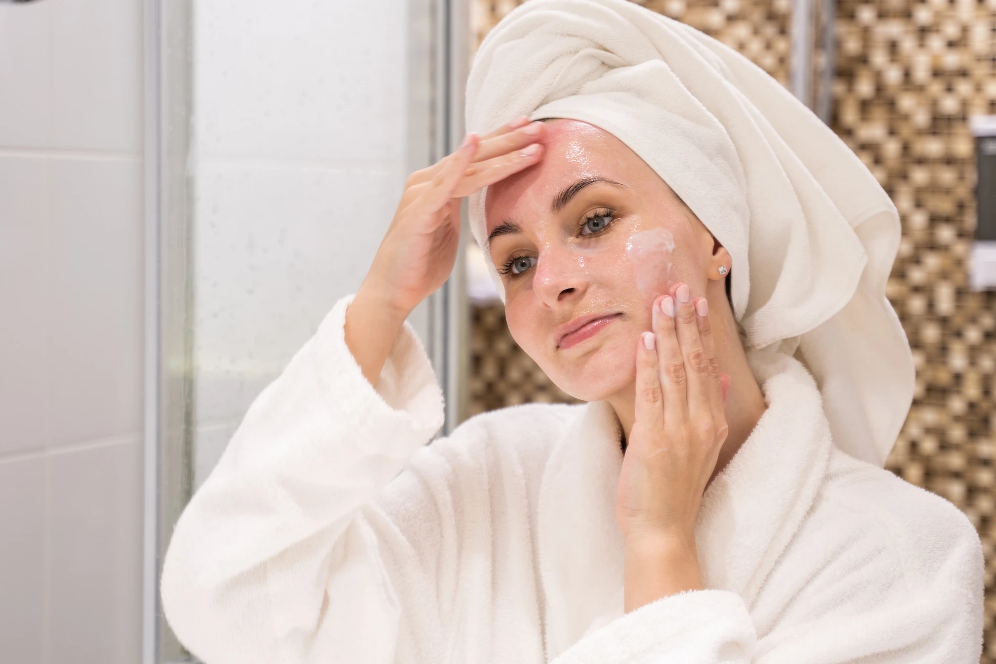 Frau im Bademantel und Handtuchturban trägt im Badezimmer eine Gesichtscreme auf ihre Haut auf